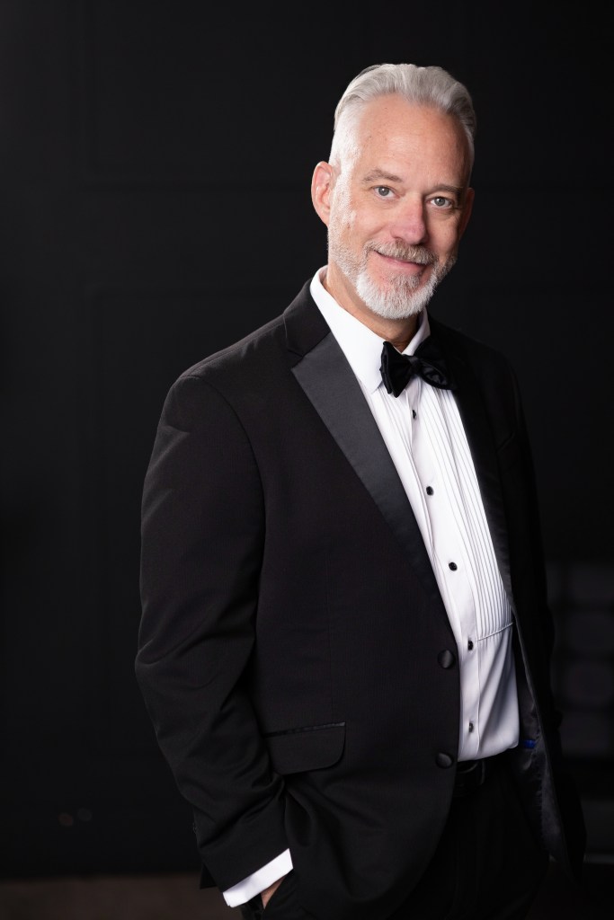 A smiling man with gray hair and a beard wearing a black tuxedo with a bow tie, posing against a dark background.