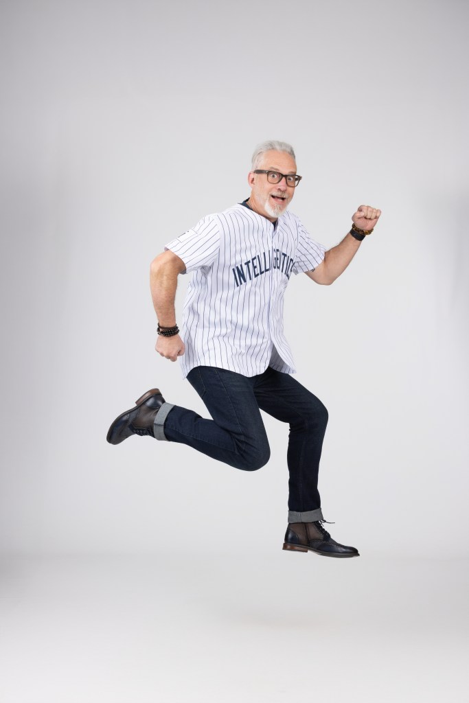 An energetic older man wearing a striped baseball jersey with 'INTELLIGENT' printed on it, jumping in mid-air with a joyful expression.