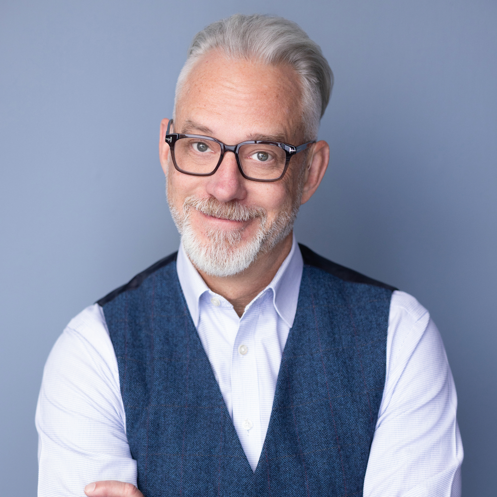 A smiling older man with a grey beard and glasses, wearing a blue vest over a white shirt, against a plain blue background.